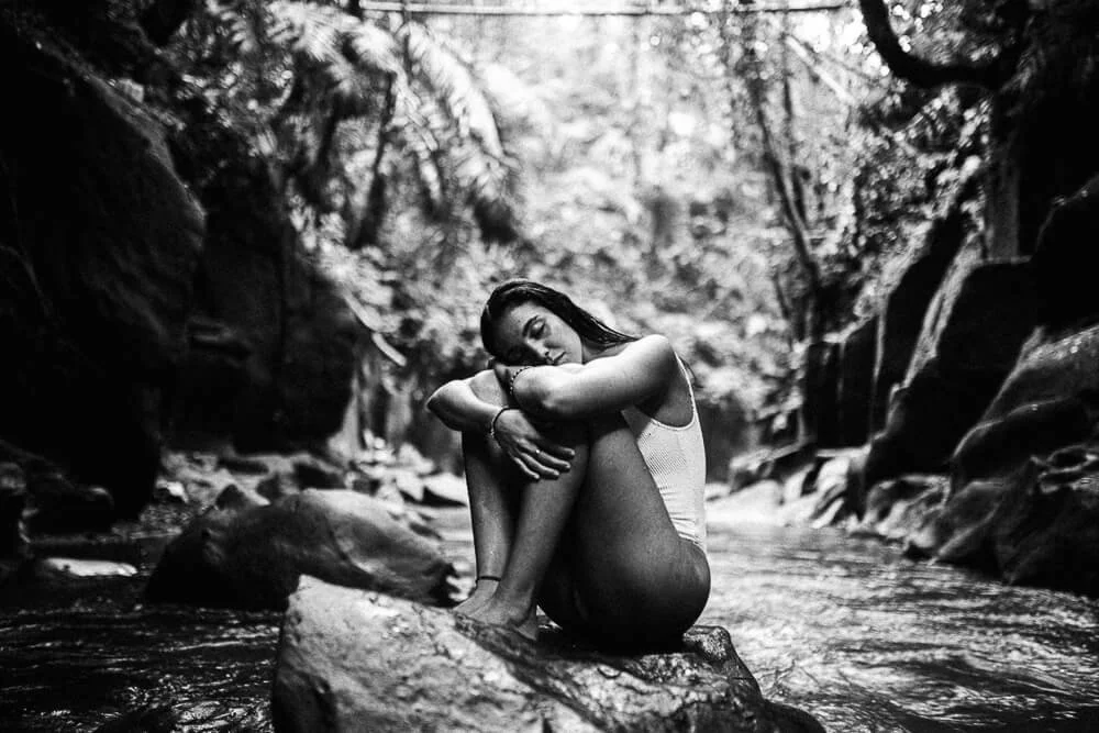 A woman sitting on a rock in a river surrounded by trees and rocks.