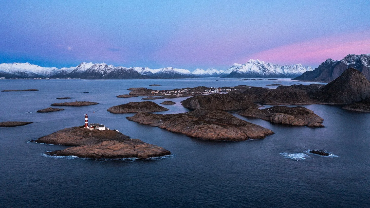 Une vue aérienne de nombreuses îles rocheuses dans une mer calme, avec un phare sur une île et un village au loin, avec des montagnes enneigées en arrière-plan au coucher du soleil.