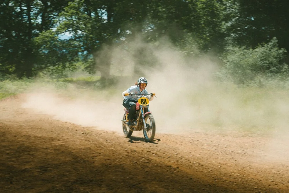 Person wearing a helmet riding a dirt bike on a wooded trail, with dust kicking up behind.