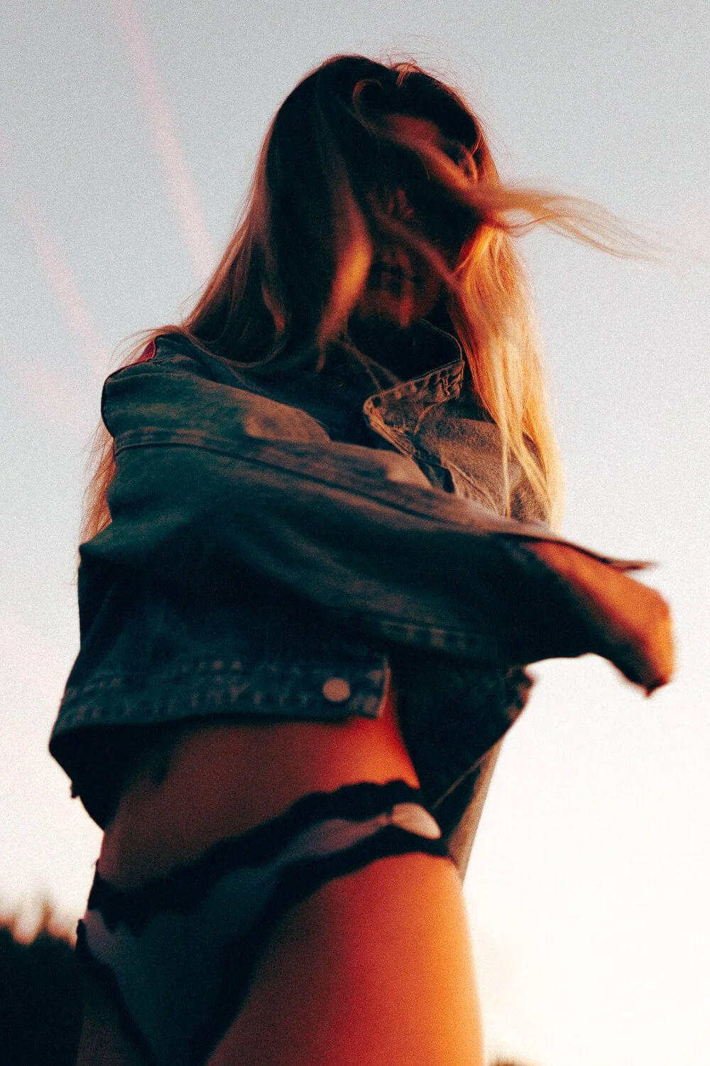 A woman with long hair is wearing a denim jacket and black underwear, standing outdoors during sunset, with her face partially covered by her hair.