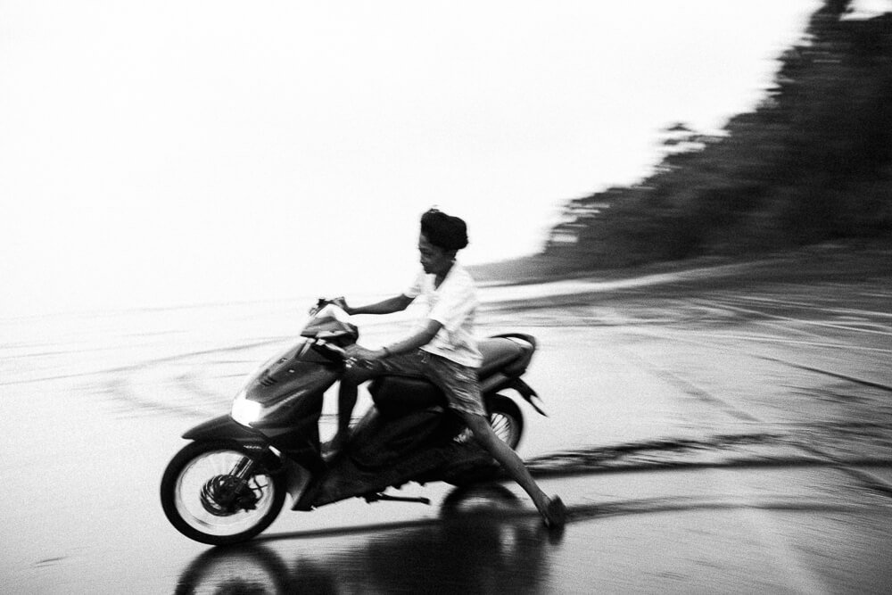 A person riding a motorcycle on a wet road, with trees on the side, taken in black and white.