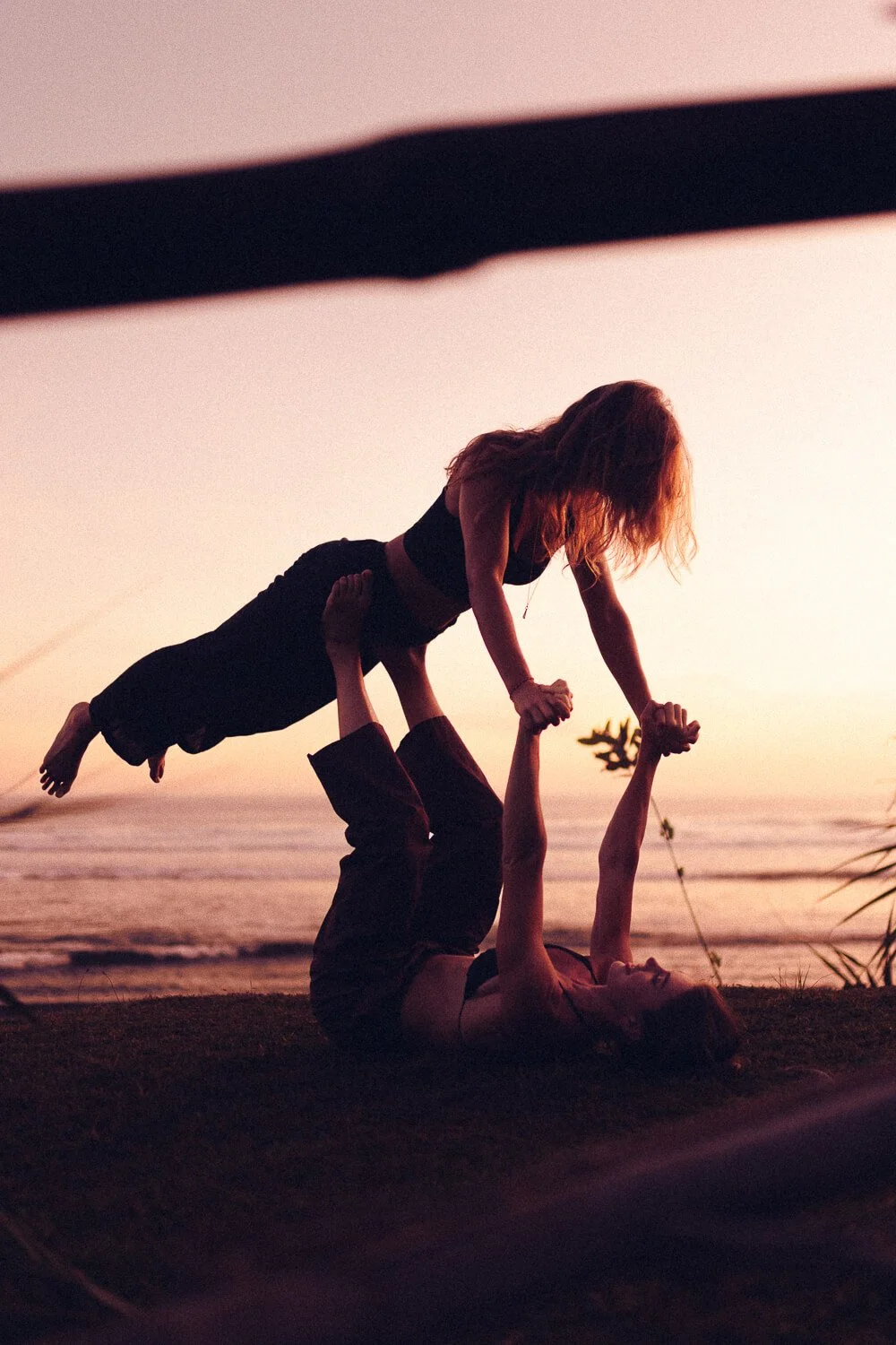 Two women performing acro yoga outdoors during sunset on the beach, with one woman lying on her back and holding the other woman above her with her legs, both holding hands.