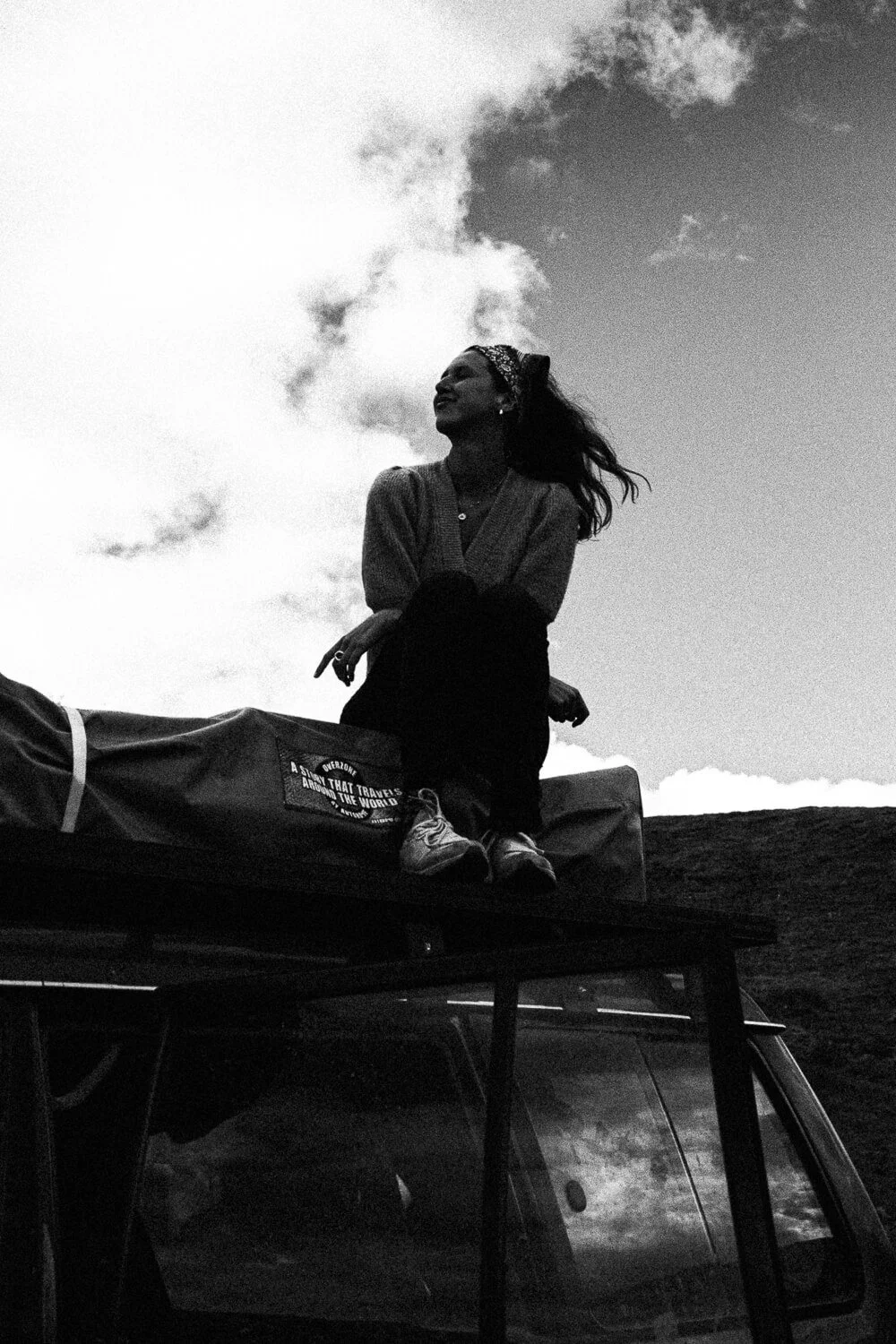 A woman with a headband and earrings sits on a roof rack of a vehicle, looking up with her hair blowing in the wind, with a cloudy sky in the background.