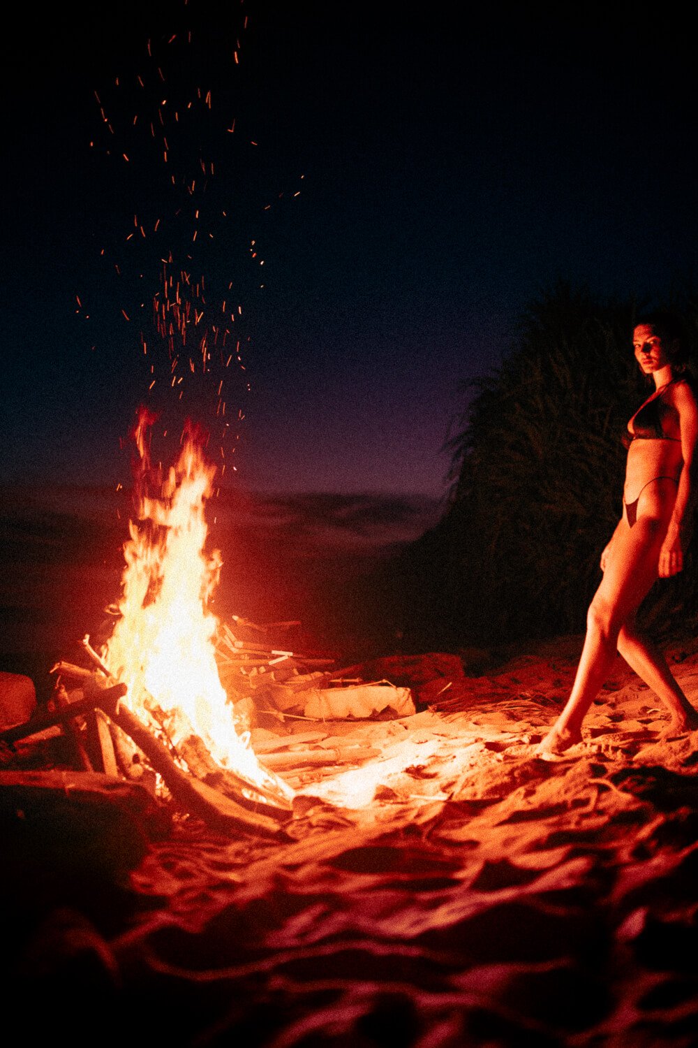 A woman in a black bikini standing on sandy beach at night near a campfire with sparks rising into the dark sky and a silhouette of a tree in the background.