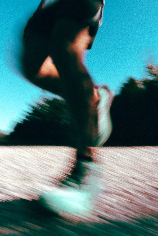 A person wearing a dark tank top and light-colored shorts, captured in motion near a body of water with a blue sky and green trees in the background, with a blurred reflection visible on the water.