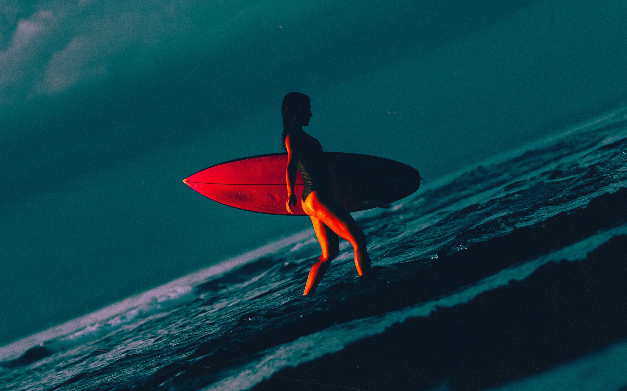Une femme silhouette portant un maillot de bain, tenant une planche de surf rouge, marchant dans l'eau de la mer au crépuscule.