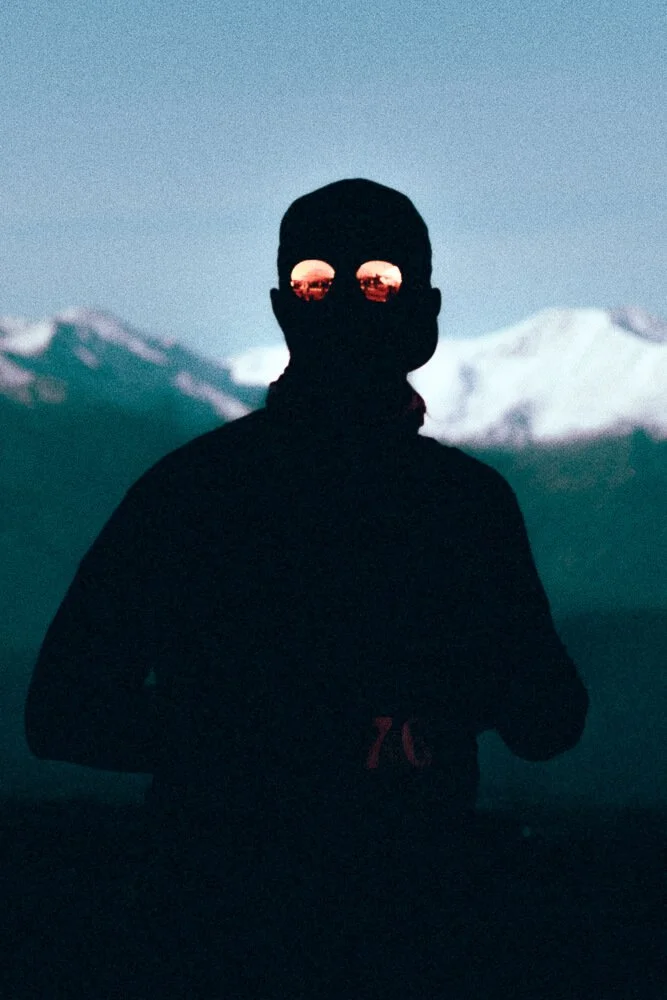 Silhouette of a person wearing a cap and sunglasses, standing outdoors with snow-covered mountains in the background during dusk or dawn.