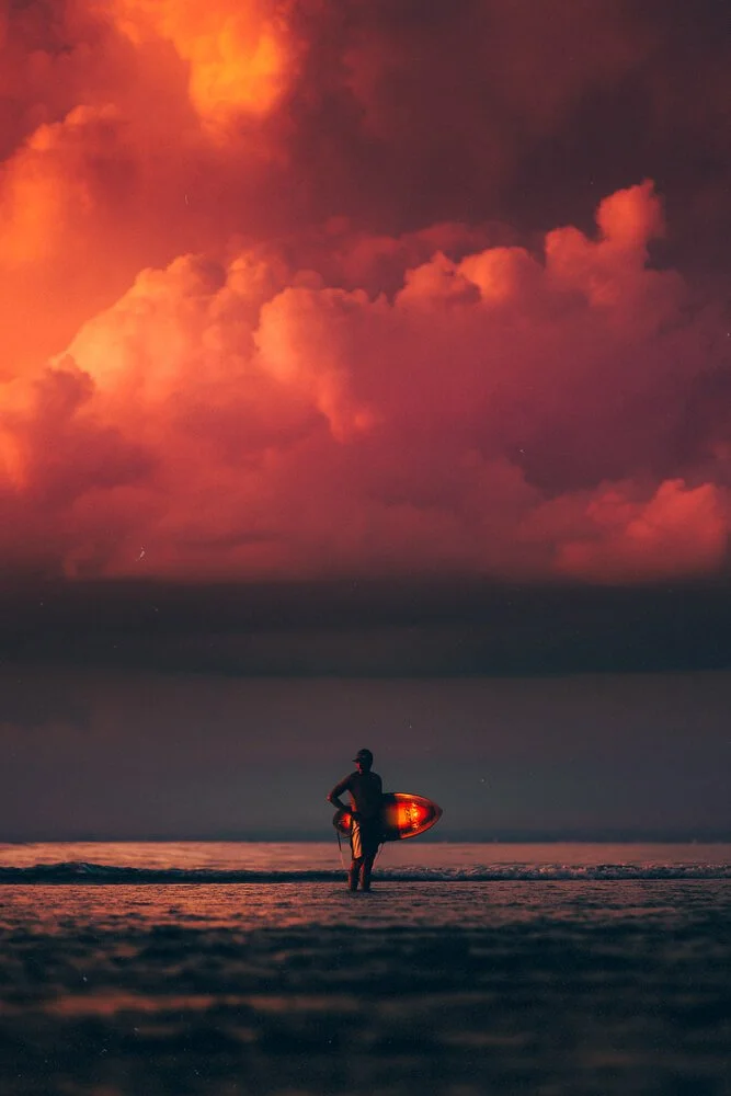A person with a surfboard walking on a beach at sunset, with a dramatic sky filled with pink and orange clouds.