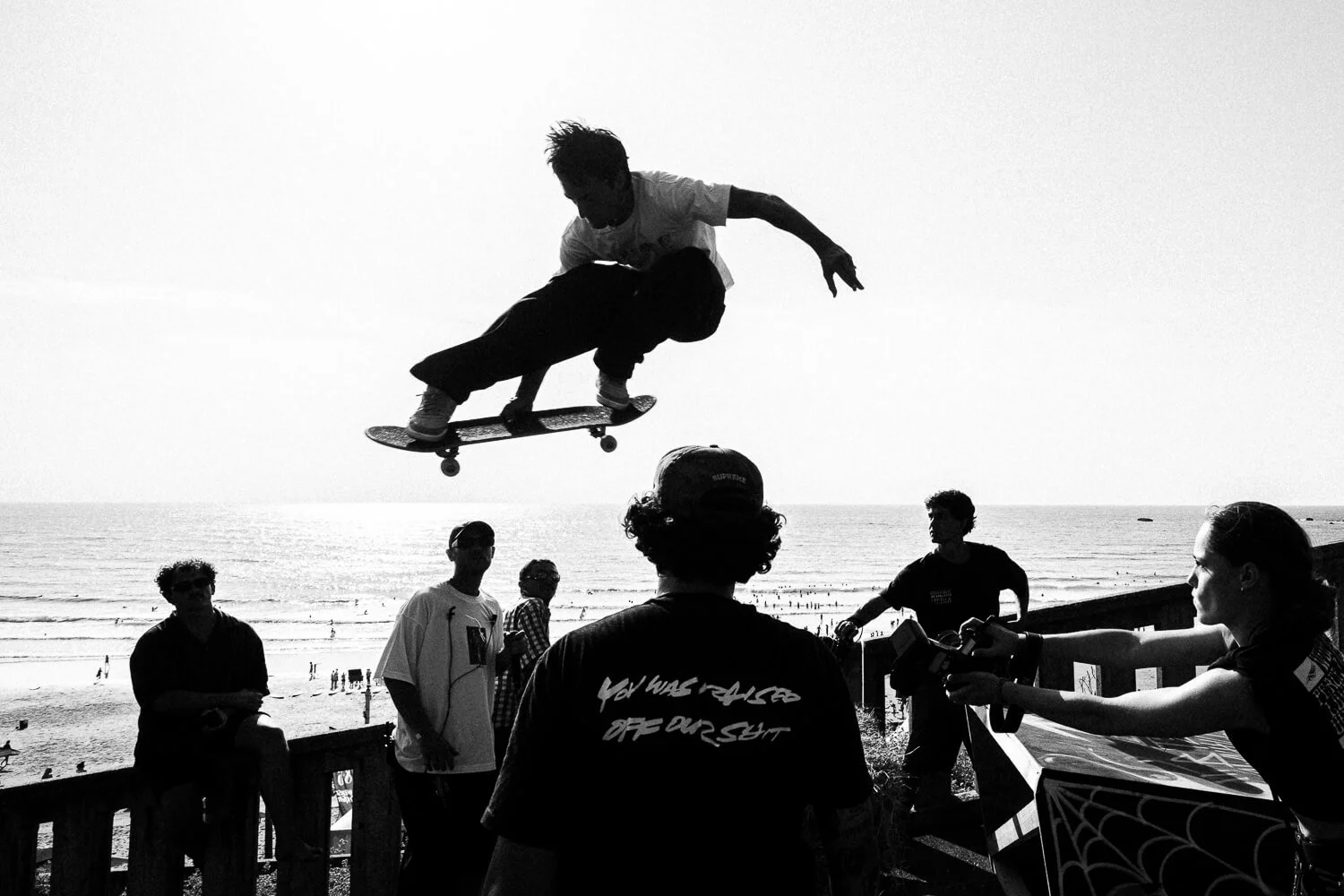 A skateboarder performing an aerial trick at the beach with onlookers and a woman taking photos.