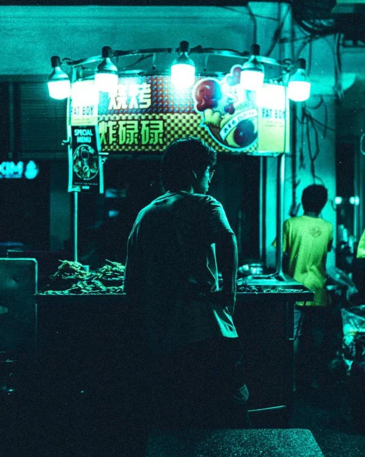 Un homme regarde un stand de nourriture ou de boissons éclairé par des lampes, dans une ambiance nocturne avec des enseignes en chinois ou japonais en arrière-plan.