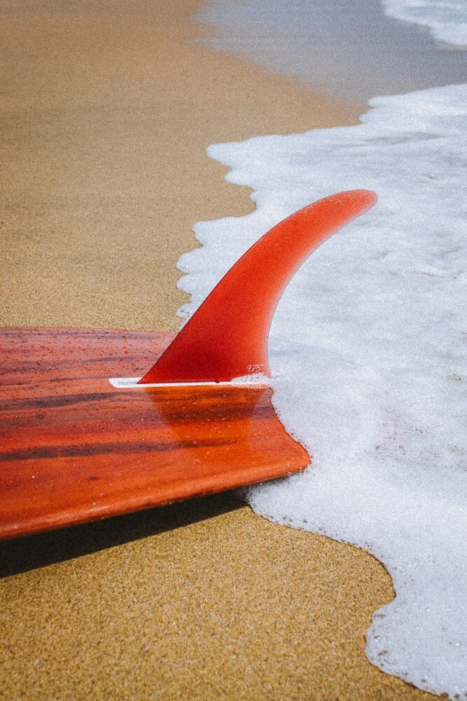 Close-up of a red surfboard with a fin, lying on wet sand at the edge of the ocean with gentle waves.