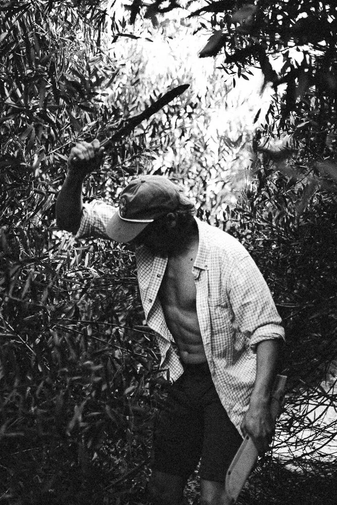 A man in a checked shirt, cap, and shorts, holding a hatchet, standing in dense vegetation or forest, with one hand on his hat, looking downward.