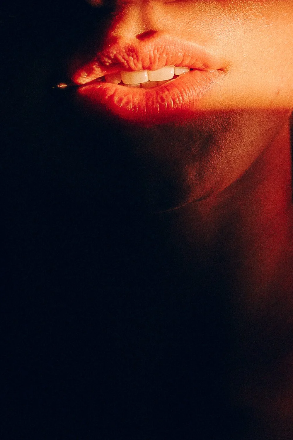 Close-up of a person's lips slightly parted, showing white teeth, with warm red and orange lighting.