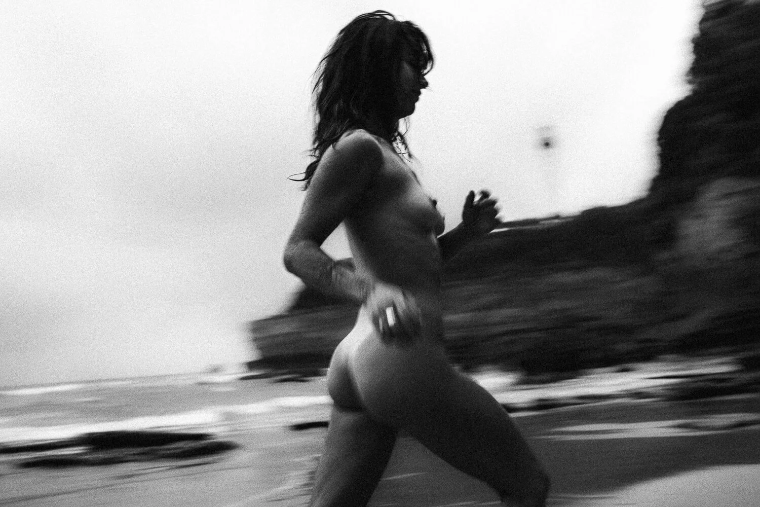 Black and white photograph of a woman running on a beach, holding a small object in her hand, with rocks and ocean in the background.