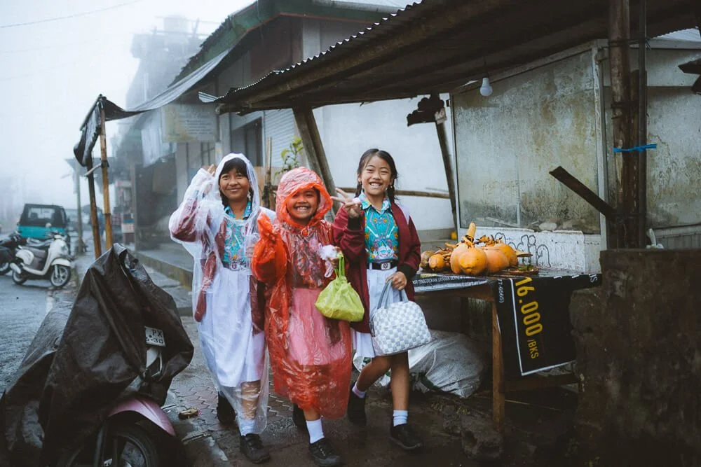 Three children in raincoats and school uniforms smiling and posing in front of a street vendor stand with pumpkins on it, in a rainy, foggy environment.
