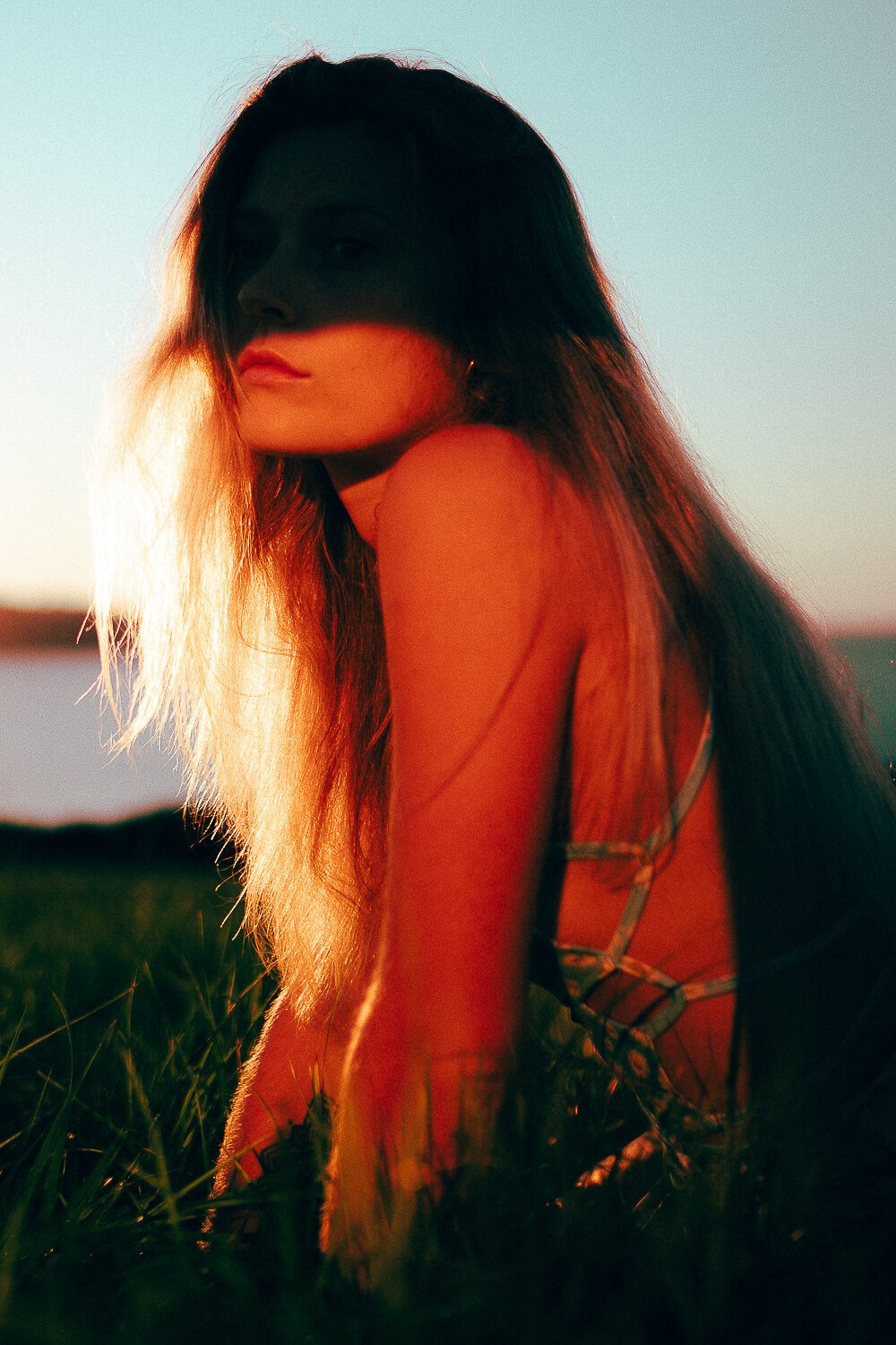 A woman with long, dark hair sitting outdoors at sunset, with her face partly shadowed, in a tank top with thin straps, surrounded by green grass and a blurred horizon.