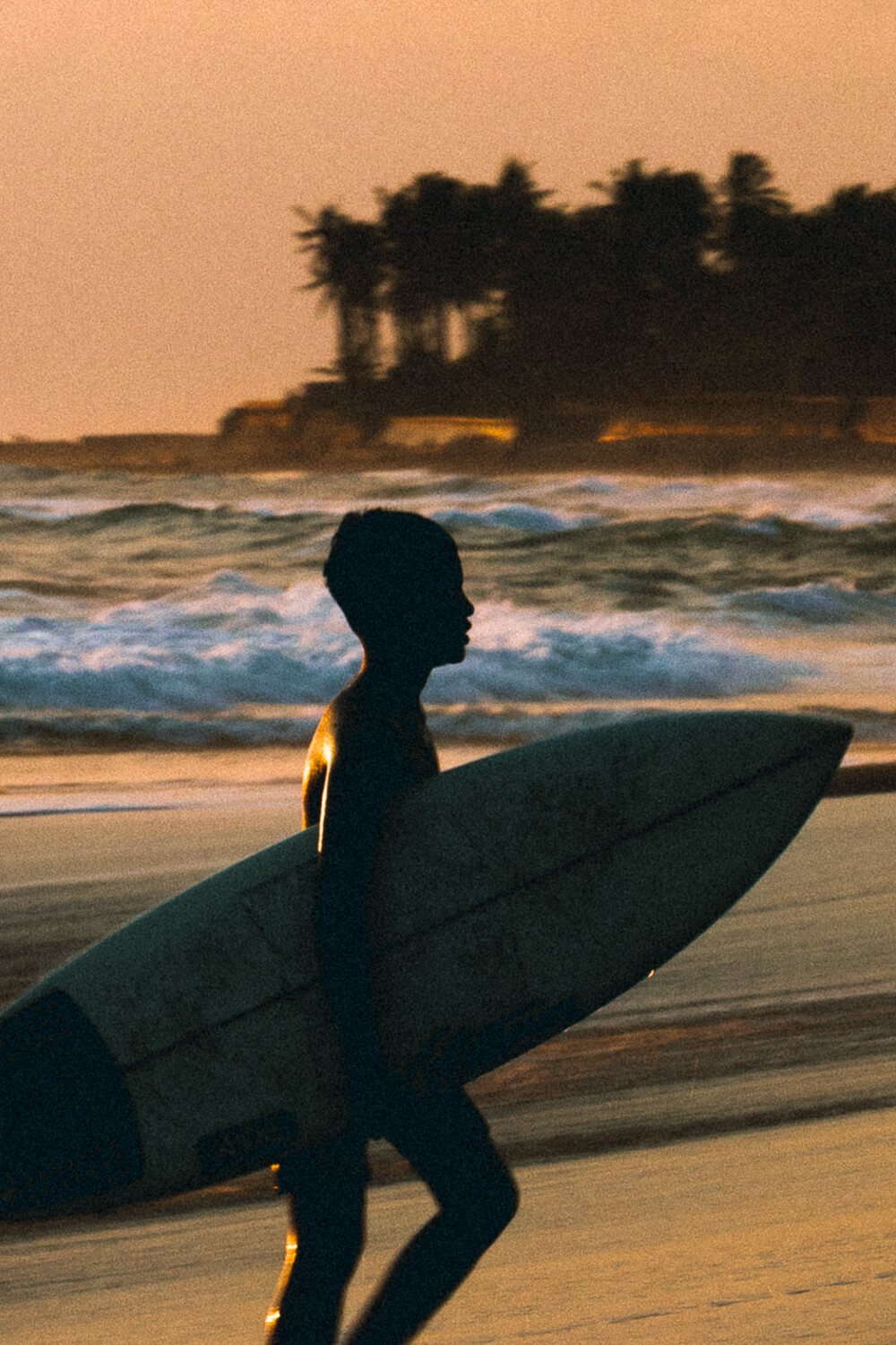 Un garçon avec une planche de surf marche sur la plage au coucher du soleil, avec des vagues en arrière-plan et des arbres sur une petite île en arrière-plan.