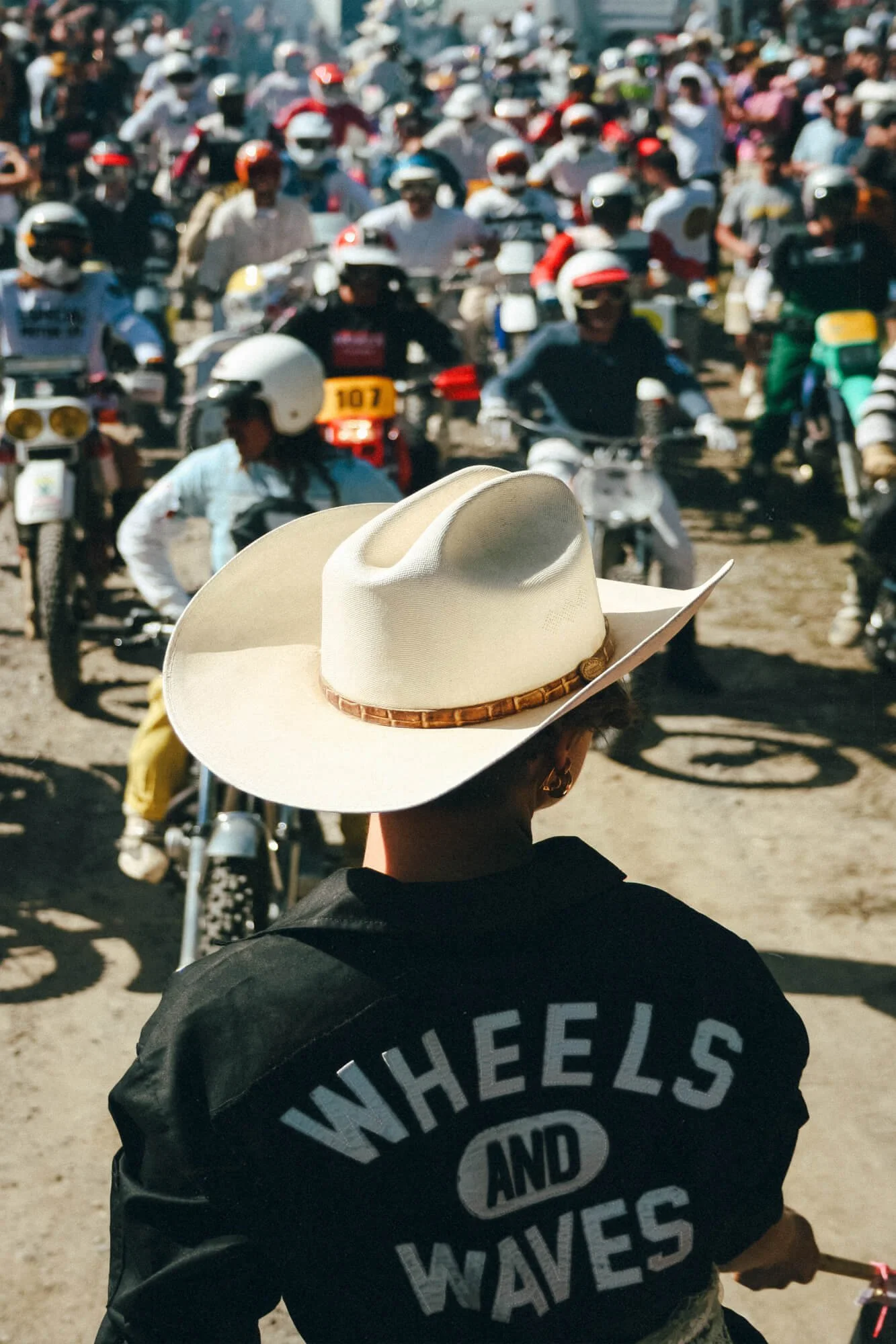 A person wearing a beige cowboy hat and a black jacket with white text that reads 'WHEELS AND WAVES' is in the foreground, facing away from the camera. In the background, there are many people riding motorcycles and scooters, wearing helmets and casu