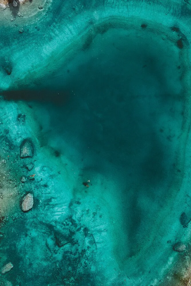 Aerial view of a blue-green geothermal hot spring with surrounding rock formations and sparse vegetation.