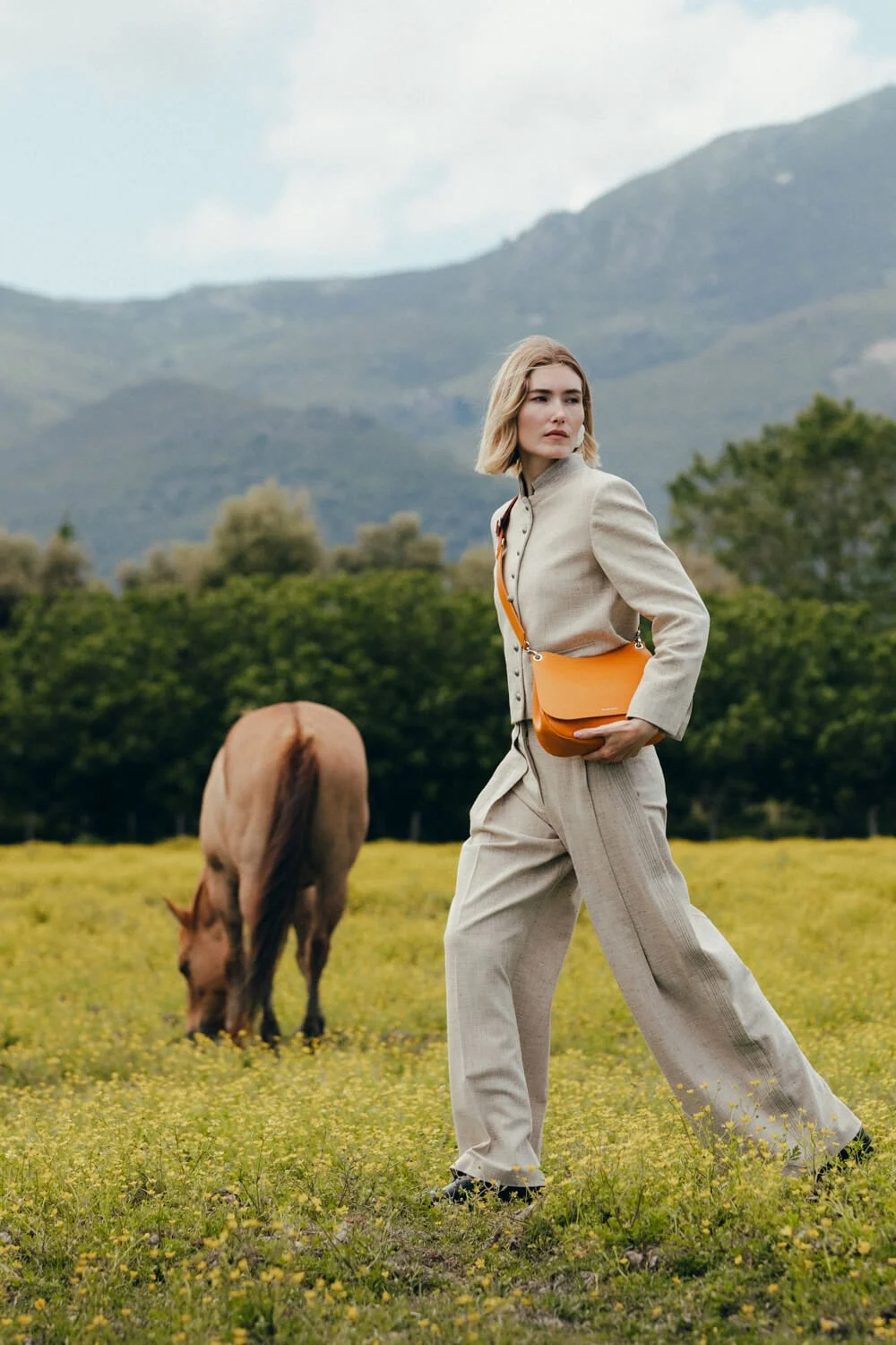 A young woman with shoulder-length blonde hair wearing a beige suit and carrying an orange purse walks through a grassy field with yellow flowers, with a horse grazing nearby and mountains in the background.