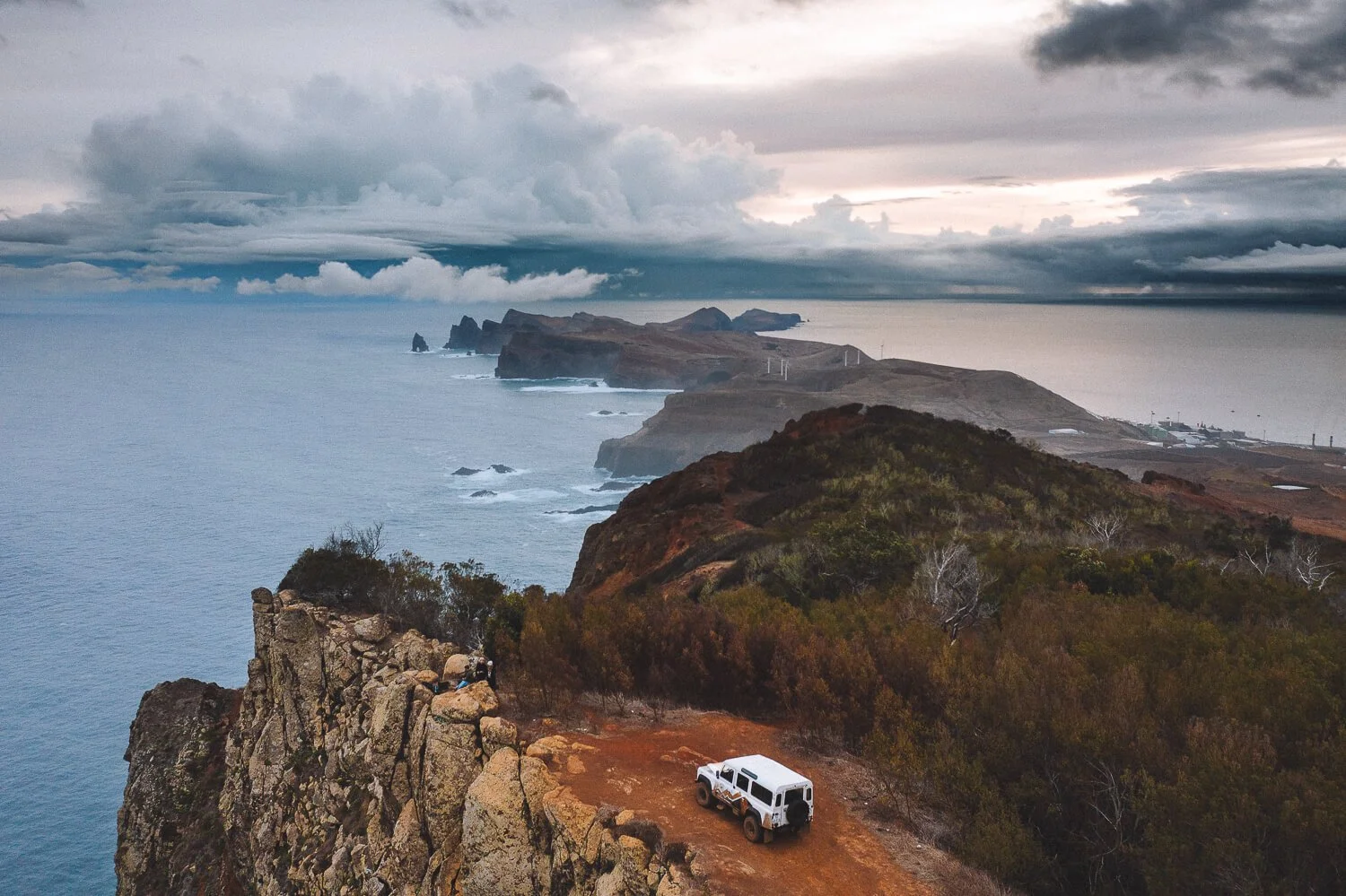 A white off-road vehicle parked on a dirt path along a rugged cliff overlooking the ocean with distant rocky islands and a cloudy sky.