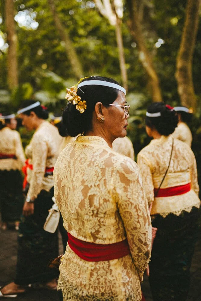Women dressed in traditional Balinese attire, featuring lace blouses, black skirts, red sashes, and white headbands, standing outdoors among trees.