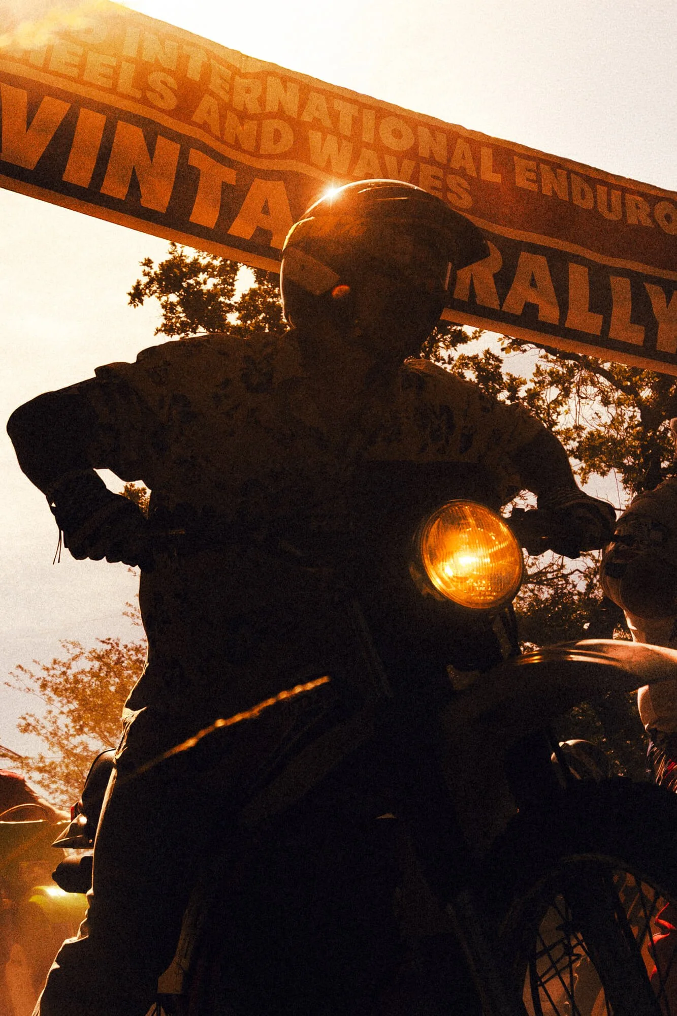 A person wearing a helmet riding a motorcycle during sunset with a banner in the background that reads "National Endurance and Wavels Rally".
