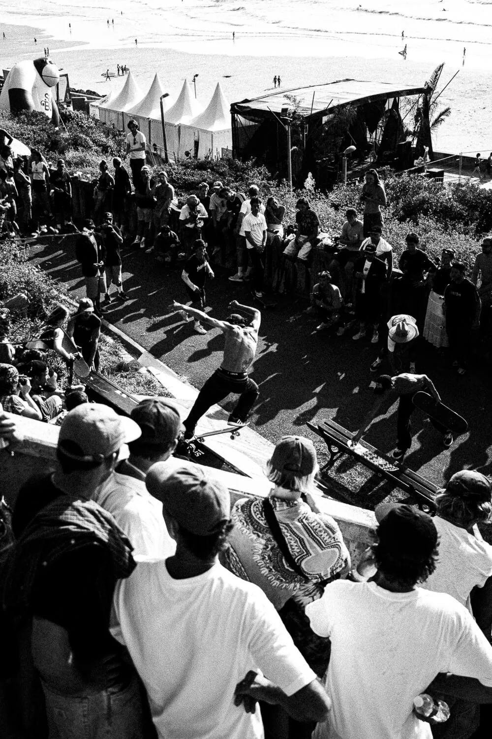 Crowd watching a skateboarder performing a trick at a beachside skatepark, with tents and the ocean in the background.