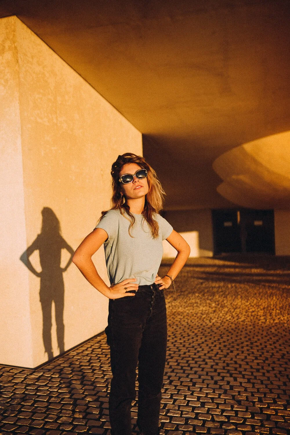 A woman standing on a cobblestone street with her hands on her hips, wearing sunglasses, a gray t-shirt, and black pants, as the sun sets casting a shadow on the wall behind her.