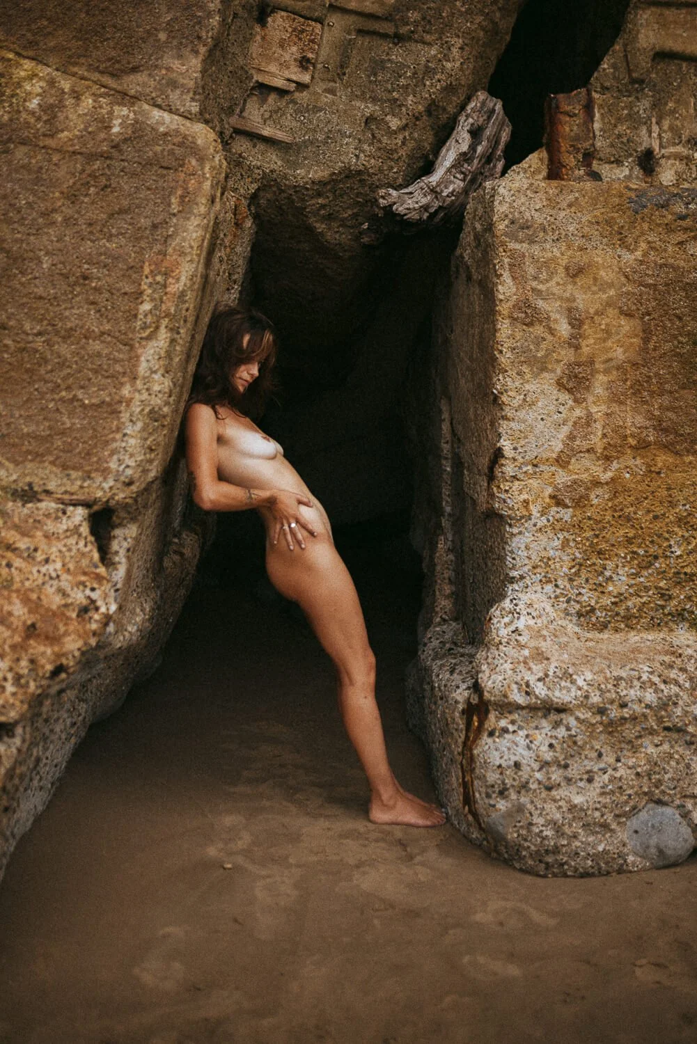A woman is standing nude, leaning against rock formations in a natural cave or rocky area, with sandy ground beneath her.