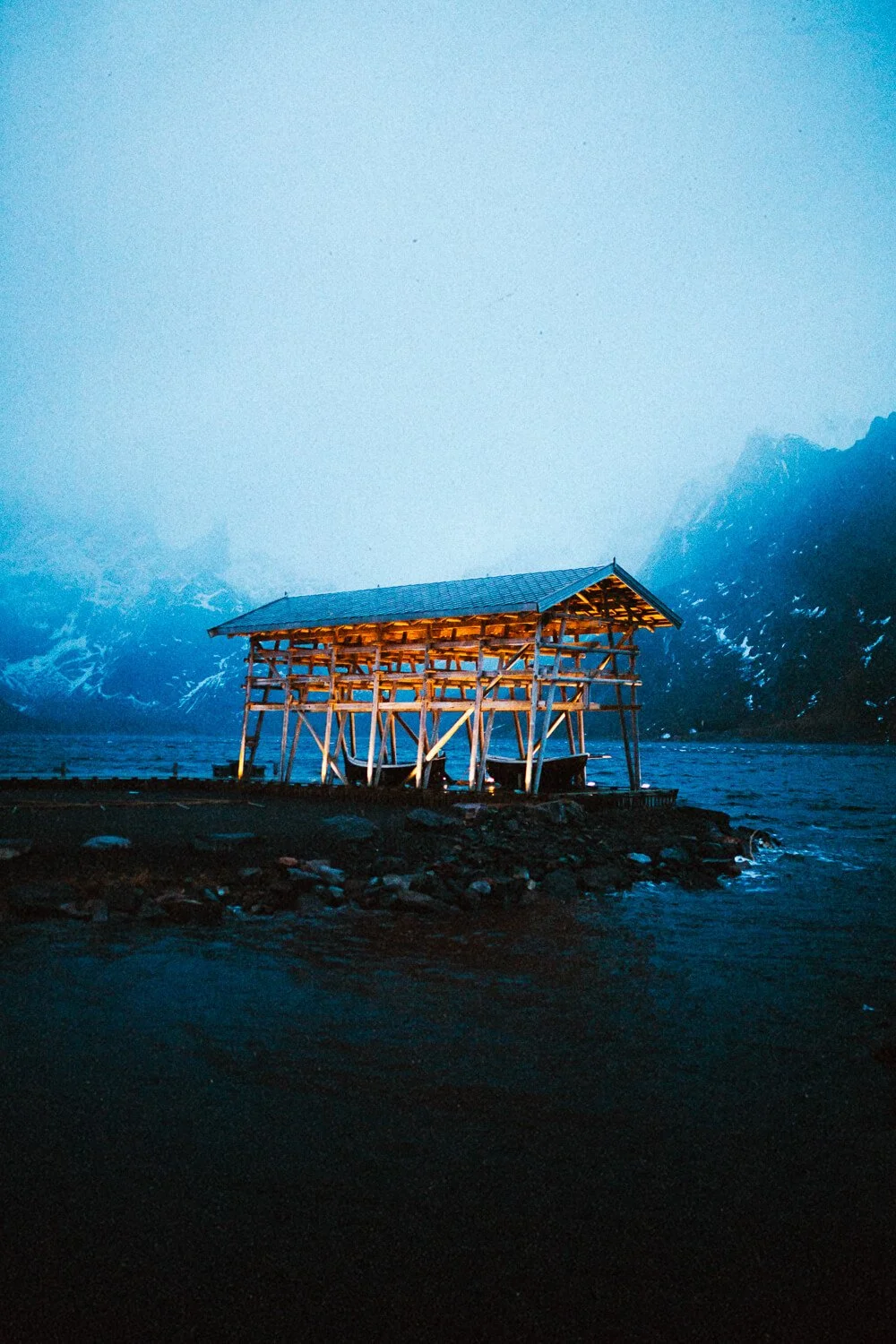 Une cabane en bois située au bord d'un lac avec des montagnes enneigées en arrière-plan, illuminée doucement dans l'obscurité