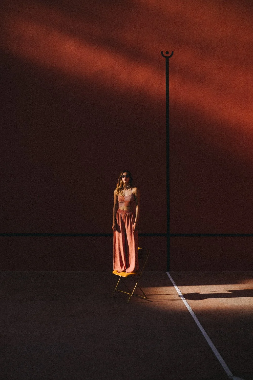 A woman with long hair wearing sunglasses, a crop top, and loose pants standing on a yellow chair, which is placed on a tennis court with a dark background and a tall pole.