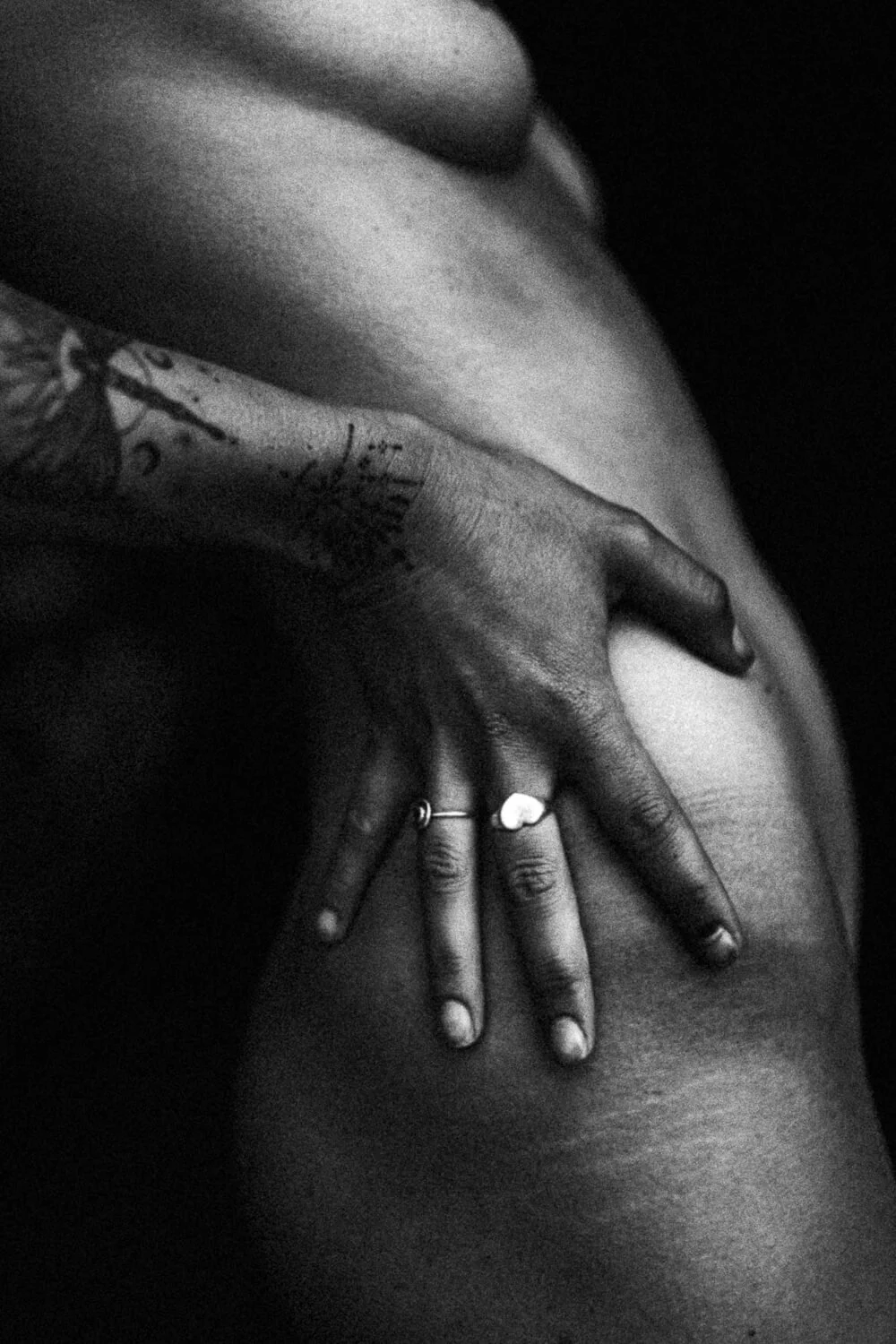 Close-up of a person's hand resting on their hip, displaying rings and tattoos, in black and white.