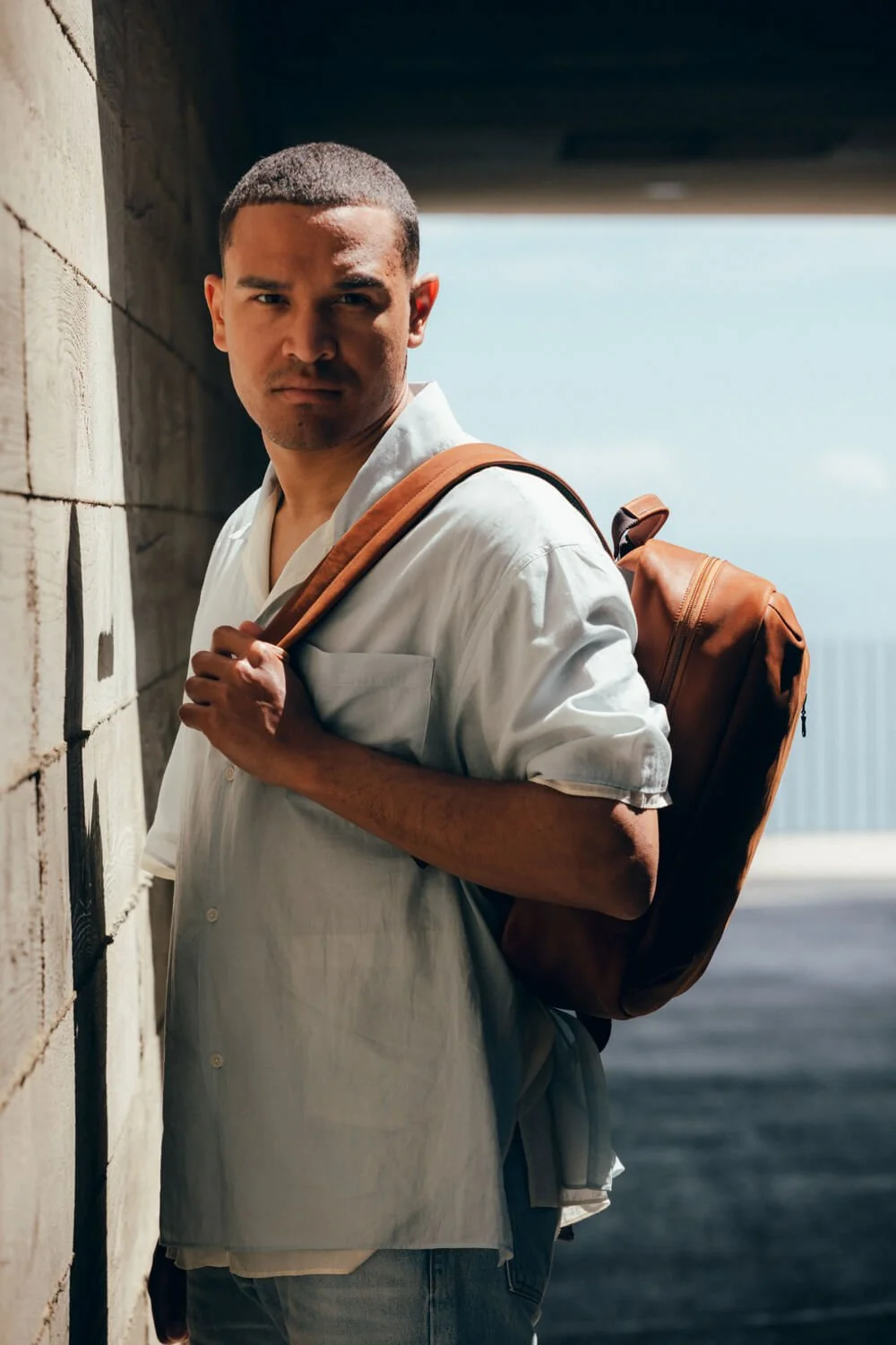 A young man with short dark hair, wearing a white shirt and carrying a brown backpack, standing outdoors near a brick wall with a blue sky in the background.