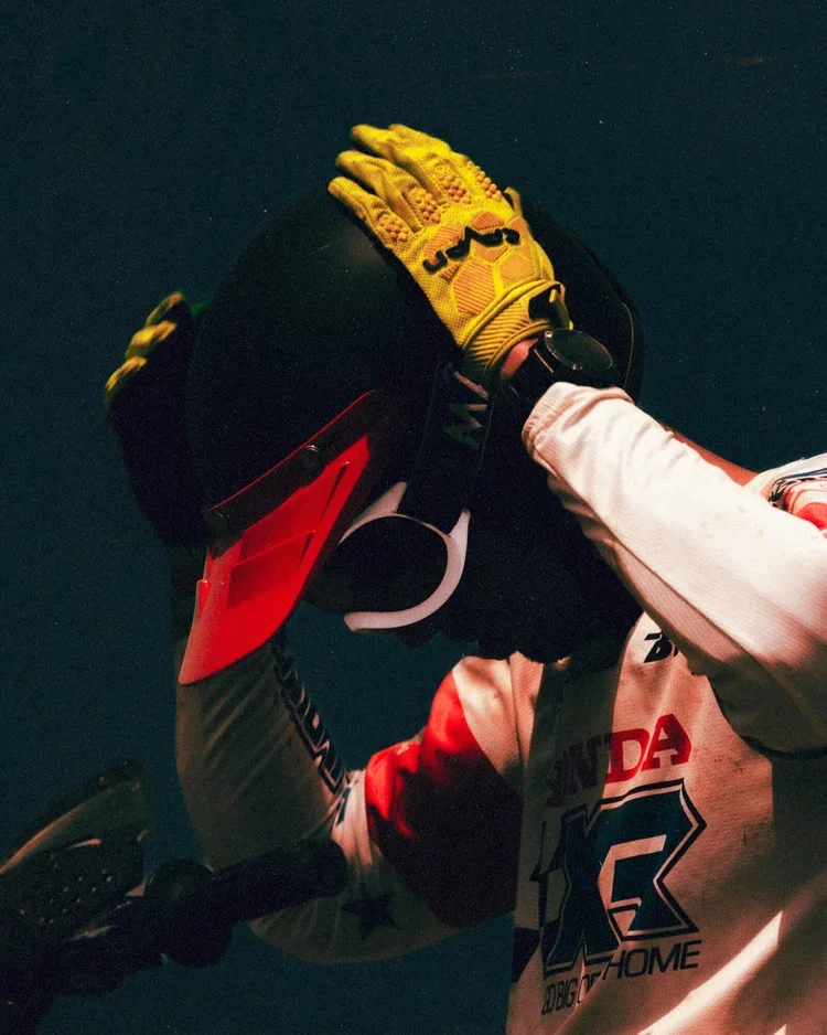 Motocross rider wearing a helmet and gloves in mid-air during a jump, dressed in racing gear with Honda and American flags logos.