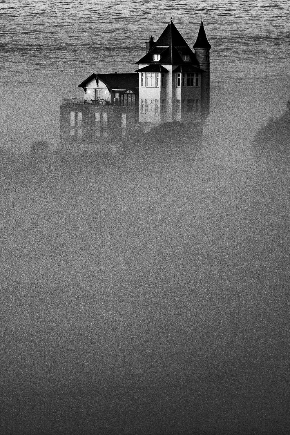 A black-and-white photo of a castle-like house on a hilltop, with fog surrounding its base.