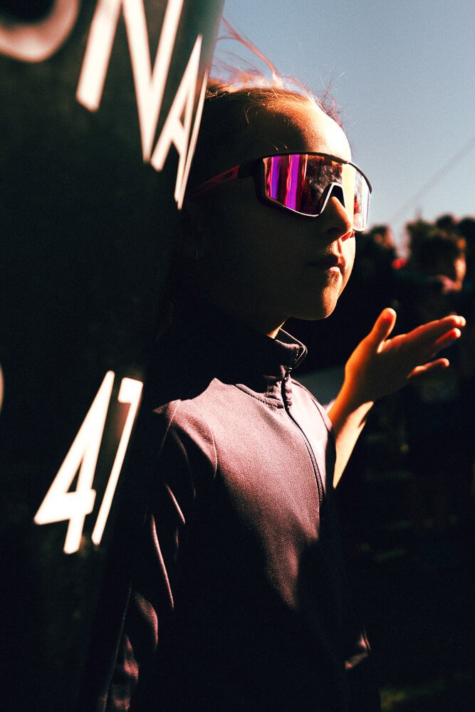 A person wearing reflective sunglasses and a purple jacket standing outdoors in the late afternoon or early evening, with a dark background and some people visible in the distance.