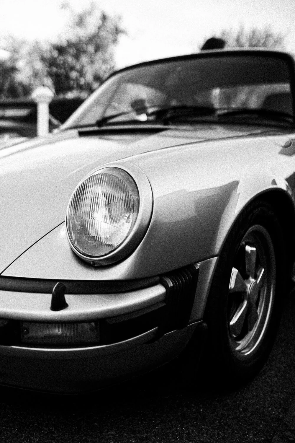 Close-up of a vintage sports car's front, showing the headlight, part of the hood, and a portion of the front wheel in black and white.