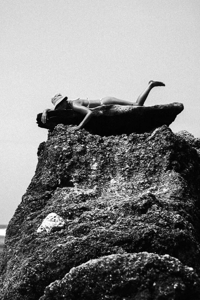 A person lying on a flat rock atop a large, textured boulder at the beach, wearing a hat and swimsuit, in black and white.
