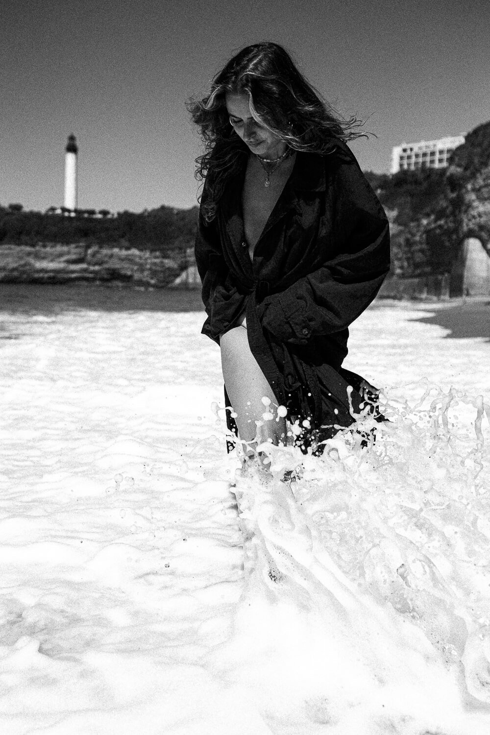 A woman with wavy hair is standing in the ocean, wearing a black jacket, with waves splashing around her.