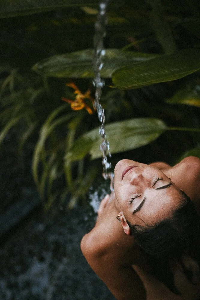 A woman with eyes closed lying underneath a stream of water falling from above, surrounded by green foliage.