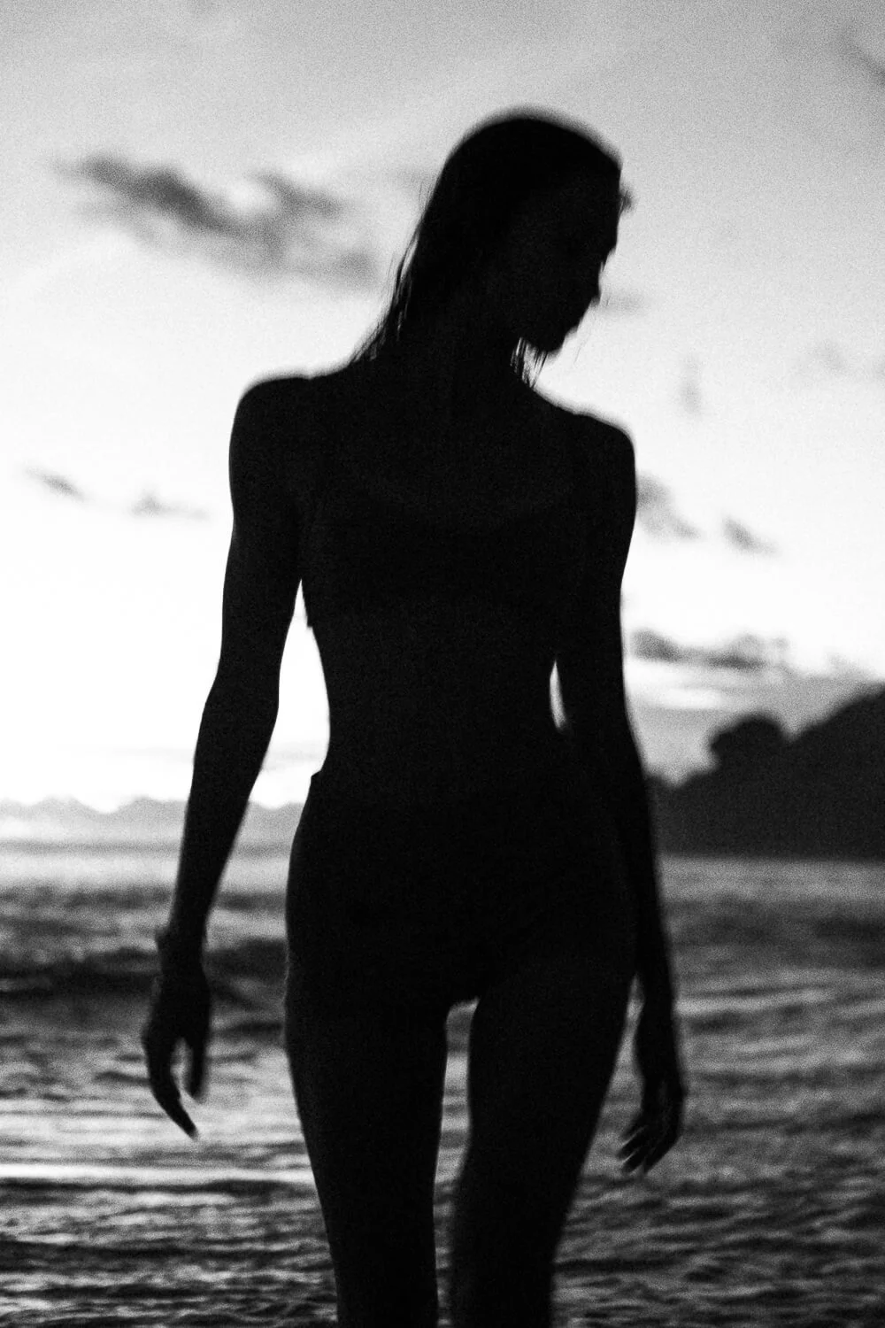 Silhouette of a woman standing at the beach during sunset or sunrise, with clouds and mountains in the background.