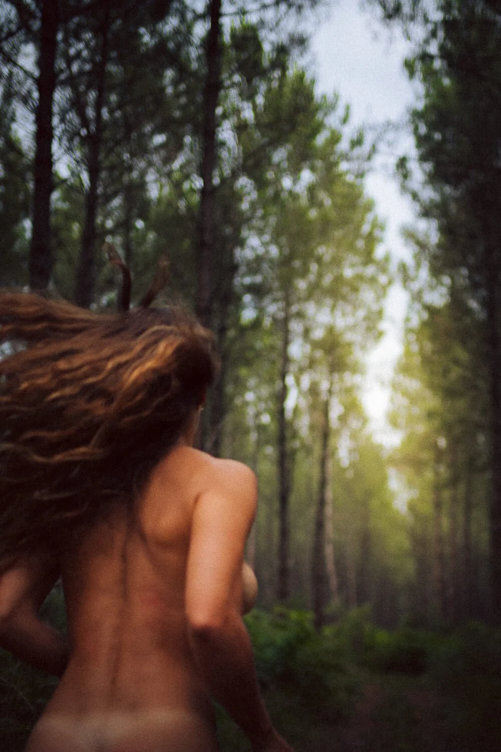 A woman with long hair running through a forest with tall trees and green foliage during daylight.