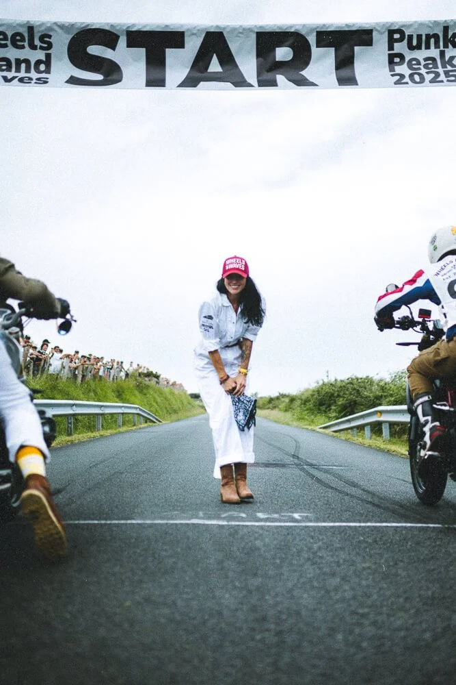 A woman in white clothing, wearing a pink cap and cowboy boots, standing on a road at the starting line of a race event, with motorcycles on either side and a large banner overhead that reads 'START' and mentions 'Punk Peak 2025'.