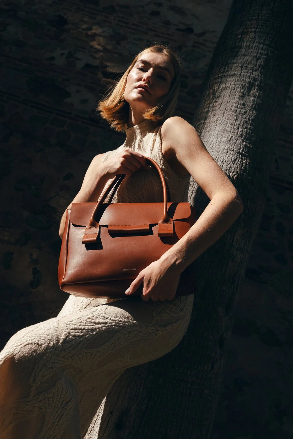 A woman holding a brown handbag, leaning against a tree in sunlight with a stone wall background.