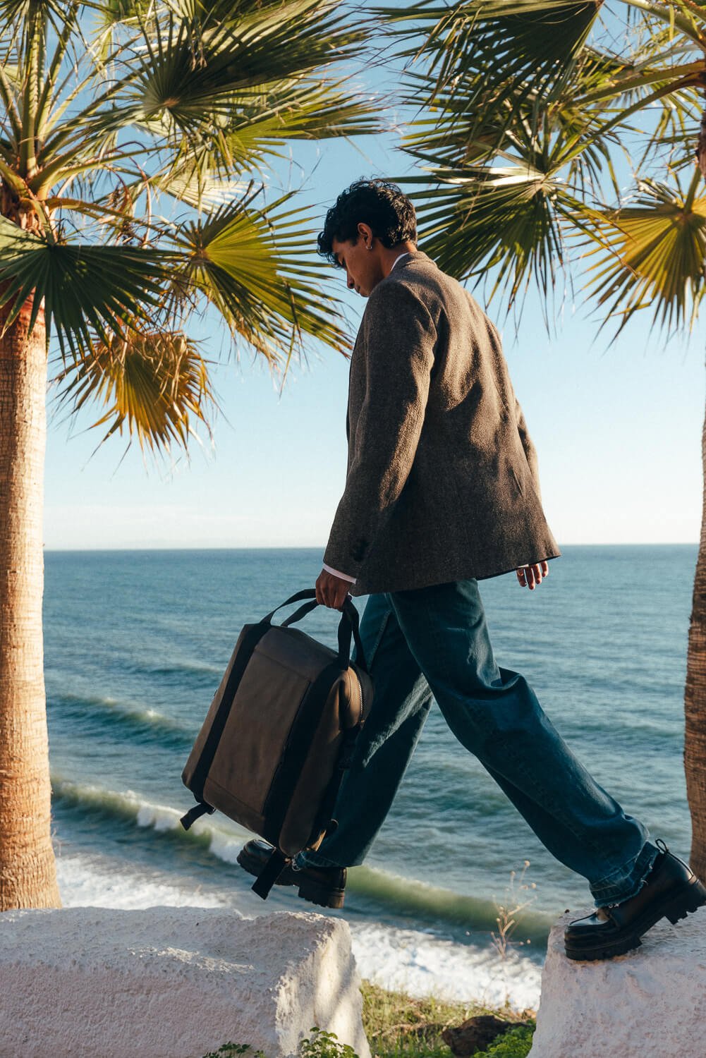 A man walking along the beach with a rolling suitcase, surrounded by palm trees, ocean waves, and clear blue sky.