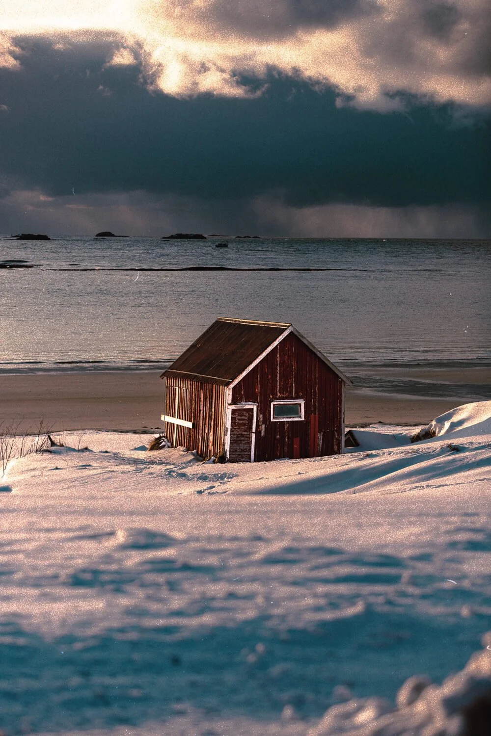 Cabane en bois rouge dans un paysage enneigé, vue sur la mer avec des nuages sombres dans le ciel.