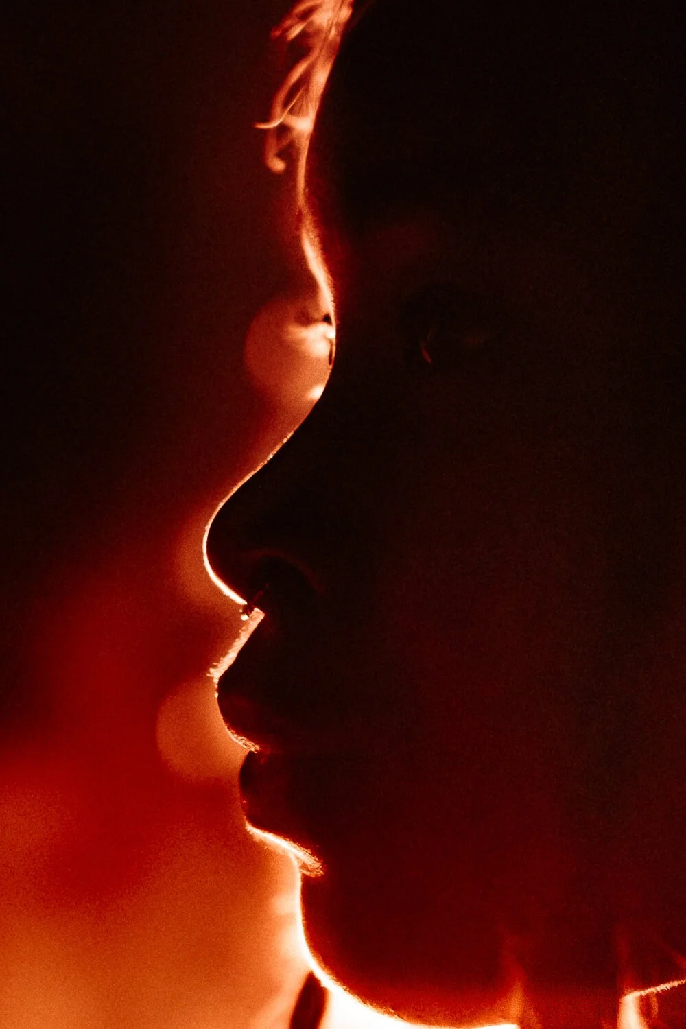 A close-up of two faces in profile, illuminated with a warm red light, appearing to be about to kiss.