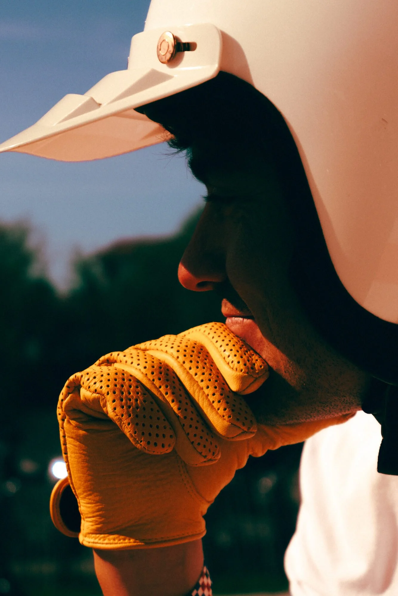 Close-up of a person wearing a white helmet and yellow gloves, holding a black object near their face, in an outdoor setting during dusk or dawn.