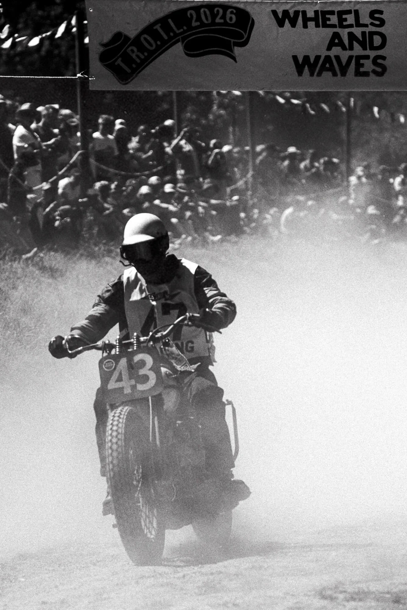 A motocross rider wearing a helmet and protective gear riding on a dirt track with a trail of dust behind. In the background, a large group of spectators and fellow riders watch the event from behind a rope, and there is a banner overhead that reads 